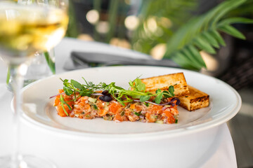 Gourmet salmon tartare with microgreens and grilled bread on a white plate, served with white wine in a fine dining setting. Elegant culinary presentation.