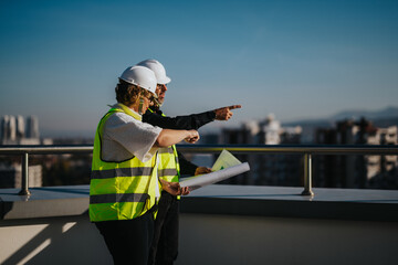 Two architects in safety vests and helmets discuss construction plans on a rooftop. They examine...