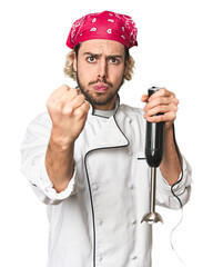 Young Caucasian chef using mixer in studio showing fist to camera, aggressive facial expression.