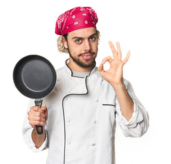 Young Caucasian male chef with frying pan cheerful and confident showing ok gesture.