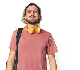 Young student with backpack and headphones blows cheeks, has tired expression. Facial expression...