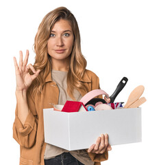 Young Caucasian blonde moving with a box cheerful and confident showing ok gesture.