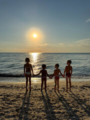 Four happy children holding hands and running into the sea at sunset on a sandy beach. Joyful summer moment, symbolizing freedom, friendship, and carefree childhood.