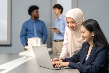 Fototapeta premium Muslim businesswoman in hijab discussing with asian colleague working on laptop in office, teamwork in a modern corporate environment, successful women collaborating on business projects, technology