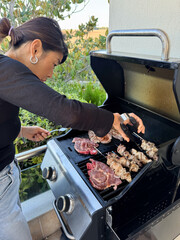 Young woman grilling meat outdoors, flipping pieces with tongs on a hot barbecue. Concept of summer cooking, outdoor lifestyle, grilling, and tasty homemade food.