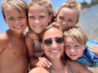 Smiling mother with her four children posing for a front-facing portrait on a sunny beach. Happy family enjoying summer vacation, love, bonding, and joyful togetherness.