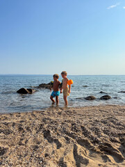 Two little kids in swimwear and armbands walking along the seashore on a sunny summer day. Concept of childhood, safety, adventure, and carefree beach vacation.