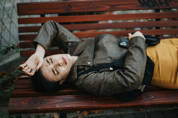 A woman lies on a wooden bench with a notable facial expression, wearing casual attire. The outdoor...