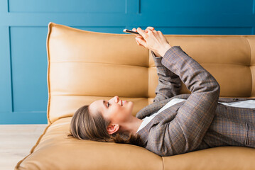 Beautiful woman with dark hair lying on the sofa in her home office, smiling and texting on her phone.
