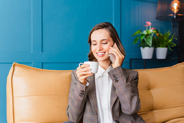 Beautiful woman with dark hair sitting on the sofa in her home office, drinking coffee, smiling and talking on her mobile phone.