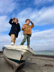 Two little boys in jackets playing on an old boat washed ashore by the sea. Atmospheric moment of childhood adventure, imagination, and outdoor exploration on a windy day.