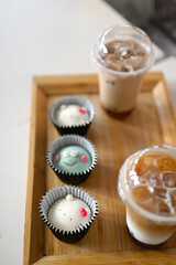 A close-up image of three adorable mochi or steamed rice cake characters arranged in black cupcake liners on a wooden tray with bubble tea in plastic cups with straws 