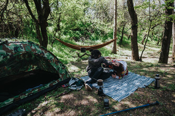 A couple bonded during camping, sitting on a blanket in a wooded area surrounded by trees, with a...