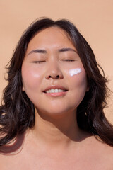 Asian woman with closed eyes, smiling gently, applying skincare cream on her cheek, showcasing a...