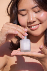 Asian woman holding a frosted glass bottle of skincare product in her hands, smiling gently, showcasing beauty and self-care in a serene outdoor setting with soft natural light