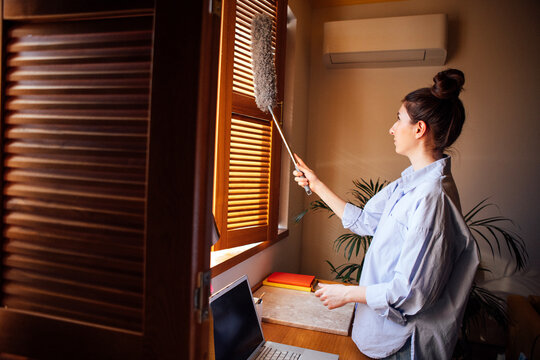 A young woman in a blue shirt is dusting wooden blinds with a pipidaster