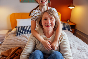 Obraz premium An elderly woman and her sweet granddaughter hug and sit on the bed. A little girl and her grandmother are smiling and relaxing in the bedroom. A mature lady and a child are having fun together