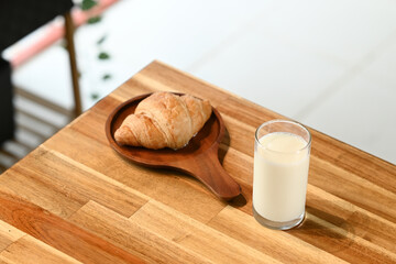 A glass of milk sits on a wooden table next to a croissant. The table is made of wood and has a brown color