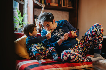 A father and child, dressed in matching seasonal pajamas, bond while sitting together indoors. The...