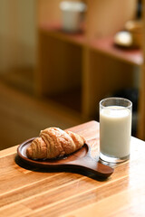 A croissant is on a wooden tray next to a glass of milk. The scene is simple and peaceful, with the focus on the breakfast items