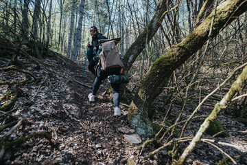A couple hiking through a dense forest, enjoying nature and outdoor exploration on a sunny day. They carry backpacks and trekking poles while navigating the rugged path surrounded by trees and moss.