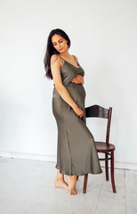 Young pregnant woman gently holds her belly. White background. Expectant mother in satin dress next...