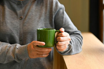 A woman is holding a green coffee cup in her hands. She is wearing a gray sweater and is sitting at a table. Concept of relaxation and comfort, as the woman enjoys her coffee in a cozy setting