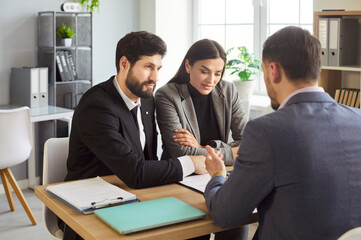 Young caucasian couple sitting at the desk discussing contract and having consultation with man realtor or financial advisor in office. Insurance agent consulting man and woman in office.