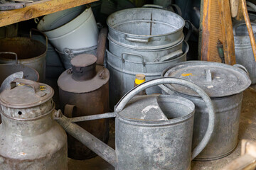 Old metal watering cans, buckets, and milk churns arranged in a rustic storage space. Visible signs of use and patina give the objects a vintage countryside look.