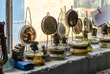 Collection of old oil lamps arranged on a windowsill. Metal reflectors visible in the background,...