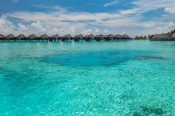  Stunning summer aerial landscape of Maldives water villas over pristine turquoise sea, luxury resort under sunny blue sky with scattered clouds, perfect tropical vacation and tourism destination