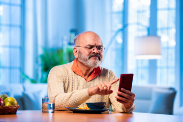 Retired Indian man browsing smartphone while sipping tea in calm morning indoor setting