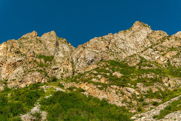 Steep rugged cliffs rise above dense vegetation, forming a dramatic landscape perfect for trekking, climbing, outdoor exploration, nature discovery, and alpine adventure in remote mountainous terrain