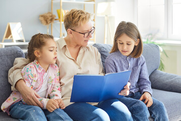 Grandmother with grandchildren reading book together during leisure at home, fostering family...