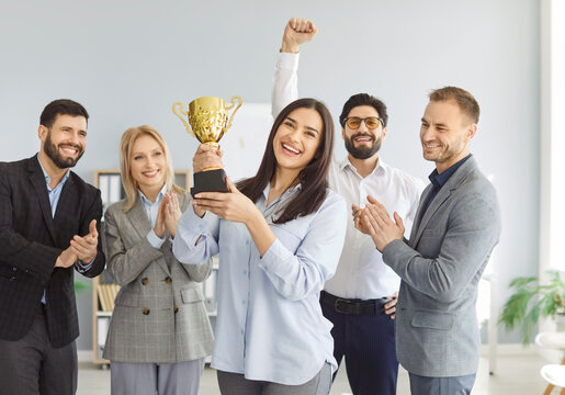 Group of cheerful business people standing in office, holding golden trophy cup and clapping hands. Happy smiling team of employees celebrating great hard work success, triumph and goals achievement.