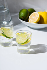 Glass cups and a jug of clean, clear drinking lemon and lime water on a white table. Sunny day and sharp shadows from glass of lemonade