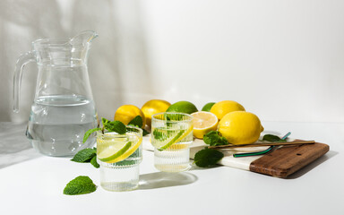 Glass cups and a jug of clean, clear drinking lemon and lime water on a white table. Sunny day and sharp shadows from glass of lemonade