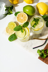 Glass cups and a jug of clean, clear drinking lemon and lime water on a white table. Sunny day and sharp shadows from glass of lemonade