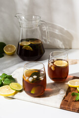 Glass cups and a jug of lemon and lime iced tea on a white table. Sunny day and sharp shadows from glass of lemonade