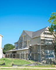 Side view new Dallas suburban home under construction. Multi-level scaffolding surrounds grey exterior, both ground and upper stories with roof completed. Grassy foreground, construction tools
