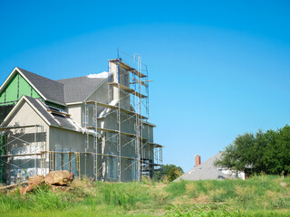 Suburban scene with incomplete home, scaffolding wraps around house foreground, finished shingle-roofed houses background. Strong contrast emphasizes architectural progression in Dallas, Texas