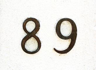 Rustic dark metal house number 89 on a lightly textured white wall. Minimalist design with aged...