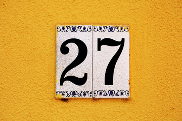Elegant ceramic house number 27 on a vivid yellow stucco wall. Decorative blue floral motifs frame bold black digits, perfect for design, address markers, Mediterranean, or European themes