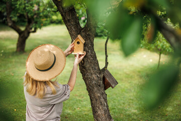 Woman in straw hat placing wooden birdhouse on apple tree in garden