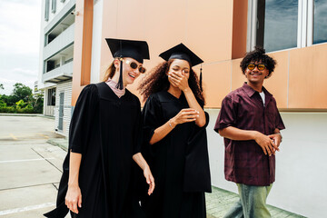 Two young women in graduation gowns and a casually dressed friend walk outside, laughing joyfully....