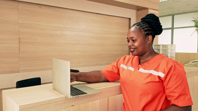 Smiling female nurse in bright orange scrubs standing at hospital reception desk with a laptop, representing healthcare staff, patient service, and hospital administration in a clinical setting.