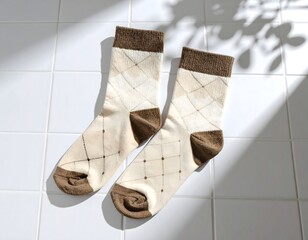 Pair of beige and brown patterned socks with diamond shapes lying flat on white tiled floor with diagonal sunlight and soft shadow from leaves