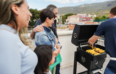 Happy friends cooking corn at barbecue rooftop dinner - Multigenerational people having fun during summer time eating and laughing together - Main focus on senior woman face