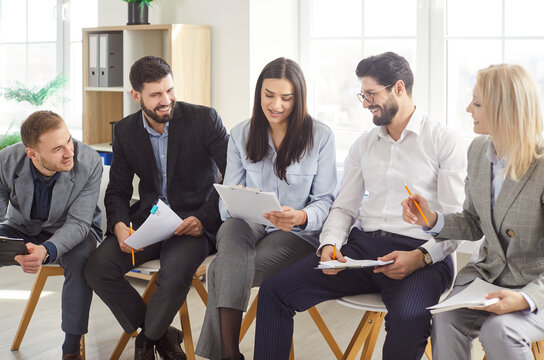 Fototapeta Group of cheerful young business people sitting in a row and talking with each other preparing for meeting or conference. Company employees chatting in office discussing new startup.