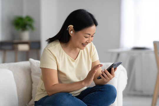A forty-year-old deaf asian  woman with a hearing aid, with short black hair, stubble, wearing a light yellow t-shirt and jeans, sits on a white fabric sofa holding a smartphone. The living room